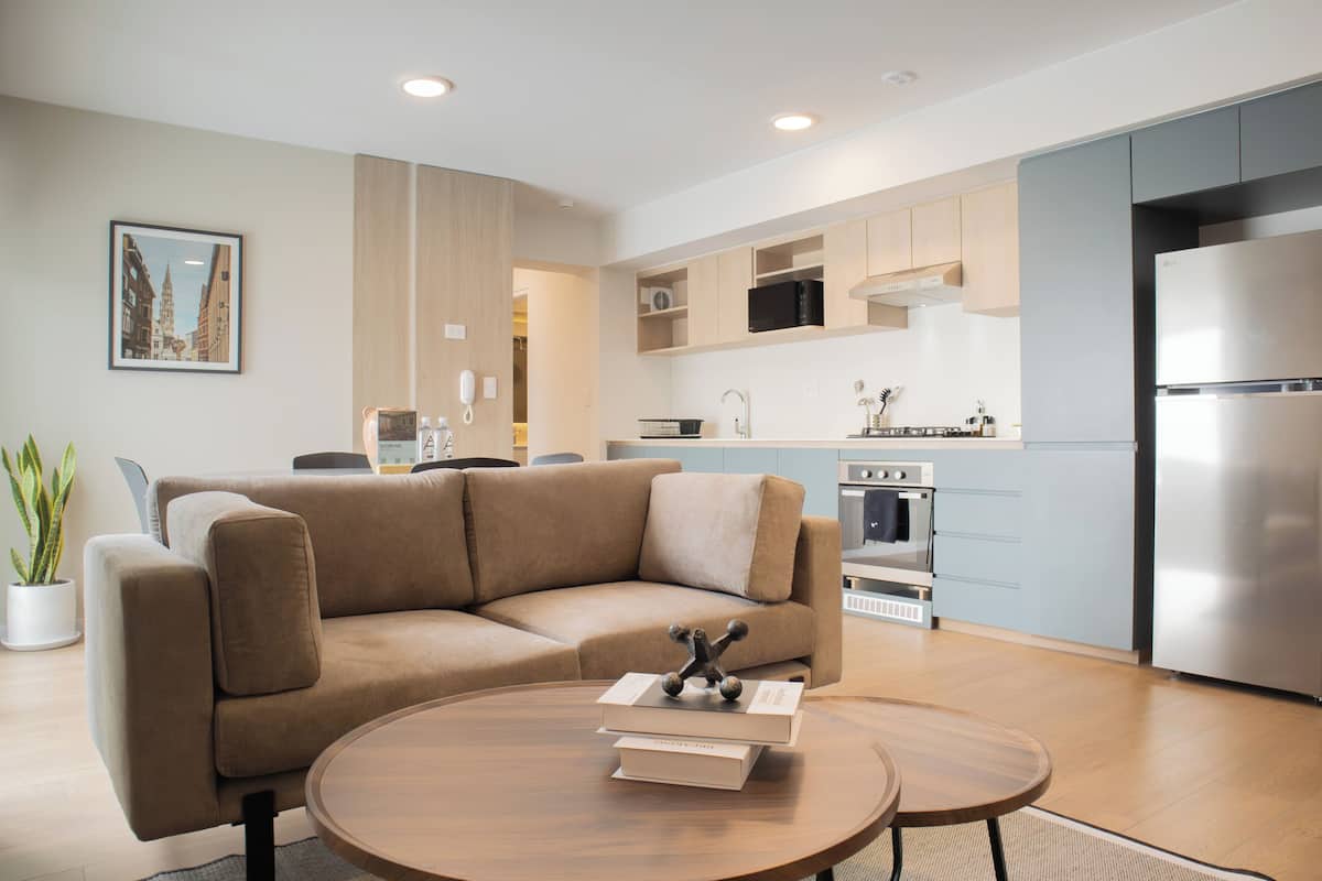 A modern living room with a beige sofa, a coffee table with books, and a kitchen area with a refrigerator, oven, and microwave.