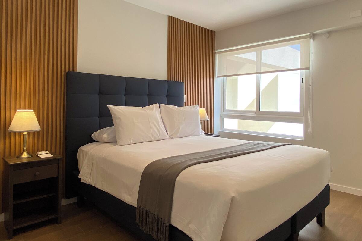 A modern hotel room with a large bed, wooden paneling on the walls, and a large window providing natural light.
