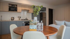 Una cocina moderna con microondas, estufa y pileta. Hay una mesa de comedor con sillas y una planta en maceta. En la mesa se encuentran un folleto y botellas de agua.