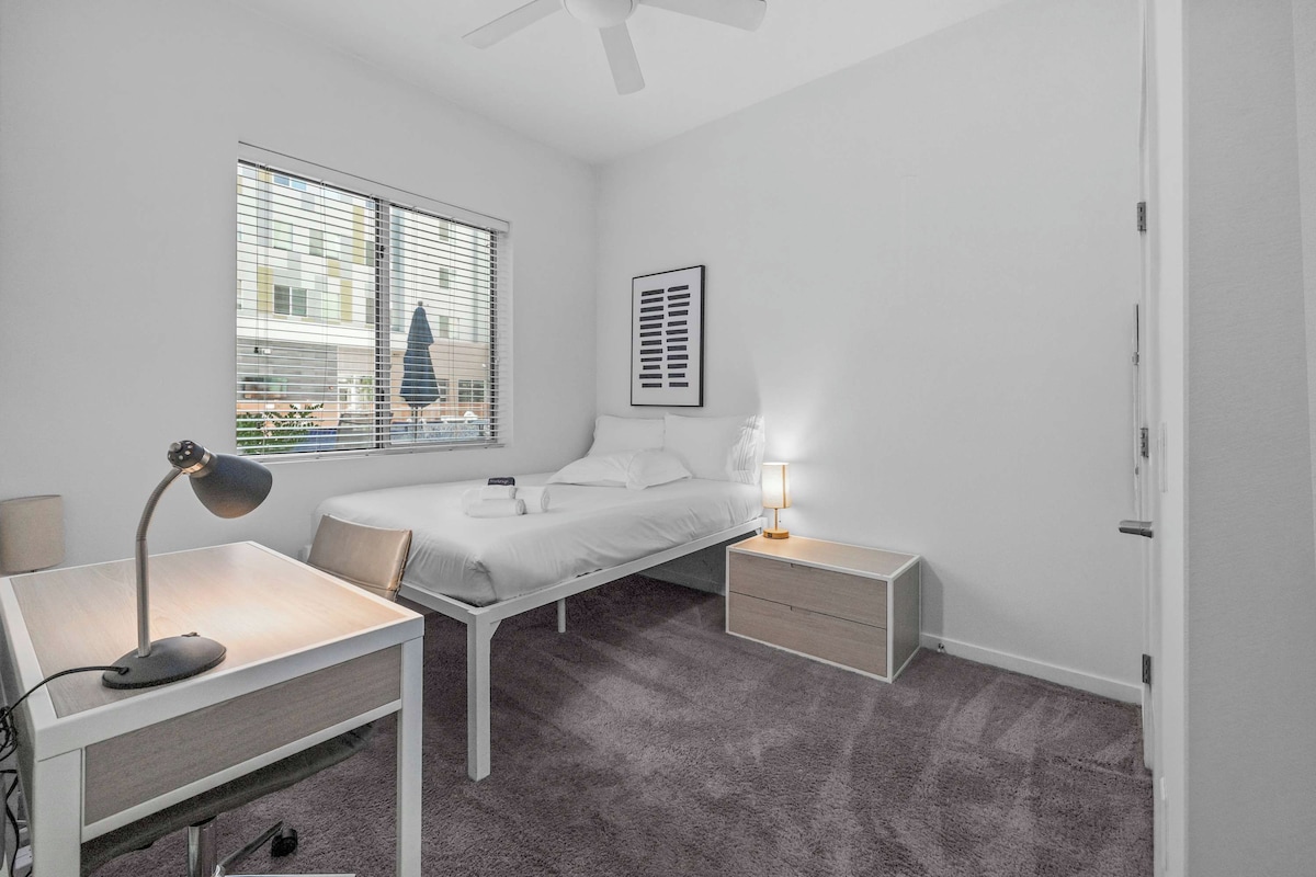 A bedroom with a bed, bedside tables, a desk, a chair, a ceiling fan, and a window with blinds.