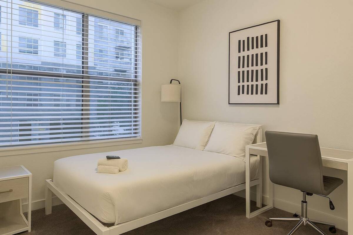 A hotel room with a bed, a desk, a chair, a bedside table, and a window with blinds.