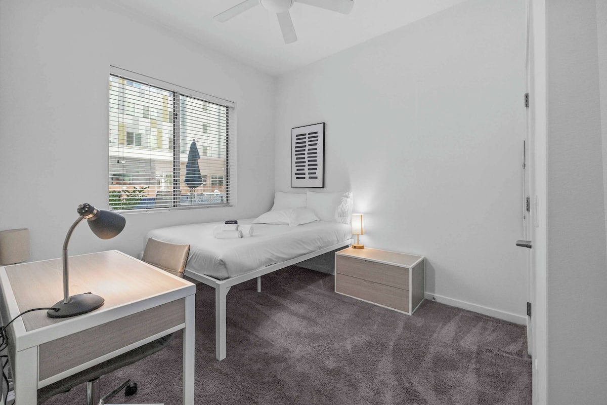 A bedroom with a bed, bedside tables, a desk, a chair, a ceiling fan, and a window with blinds.