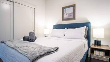A neatly made bed with a blue headboard, white bedding, and a gray blanket. There are two bedside lamps on either side of the bed.