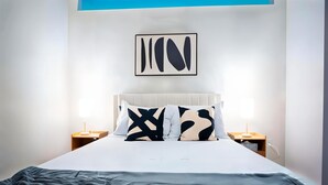 Una habitación de hotel moderna con una cama grande, un cabecero blanco y una obra de arte abstracta enmarcada sobre ella. Hay dos mesitas de noche con lámparas.