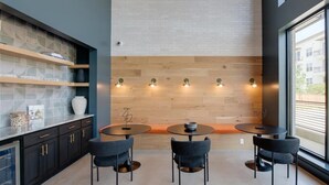 Una cocina moderna con armarios oscuros, encimera blanca y pared de madera. Hay tres mesas redondas con sillas, y un ventanal grande a la derecha.