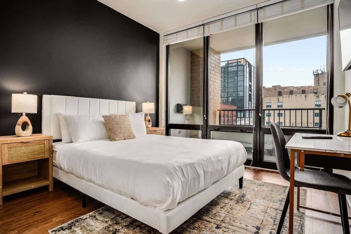 A modern hotel room with a large bed, two bedside tables with lamps, a small desk with a chair, and a large window offering a city view.