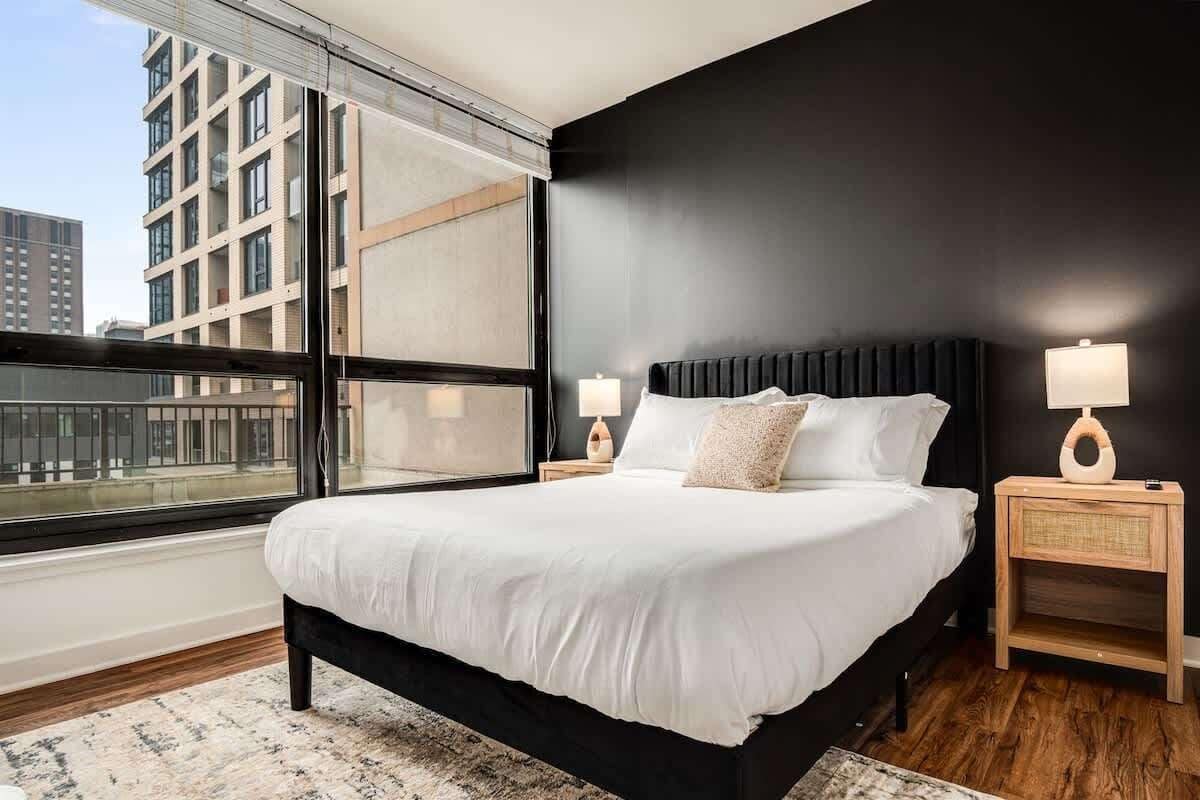 A modern bedroom with a large bed, two bedside tables with lamps, a view of the city through a large window, and a dark accent wall.