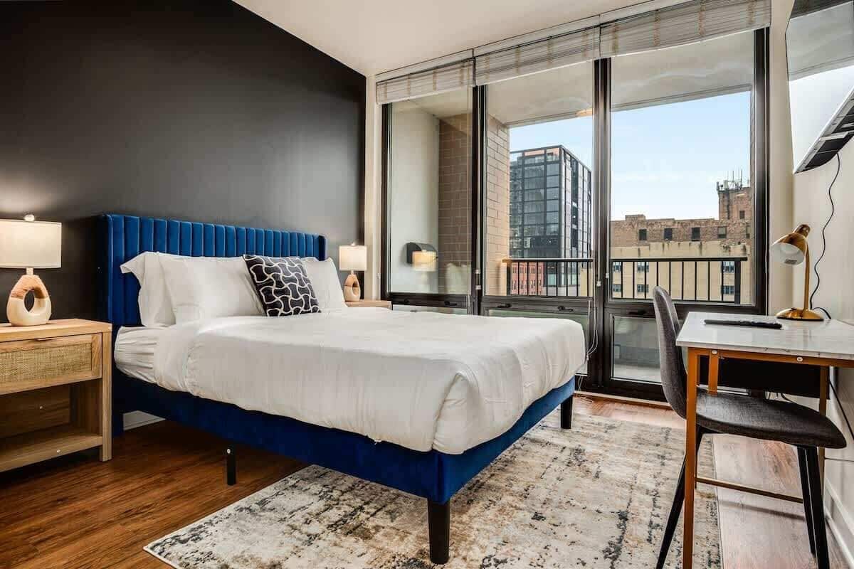 A modern hotel room with a large bed, a desk, and a view of the city through large windows.