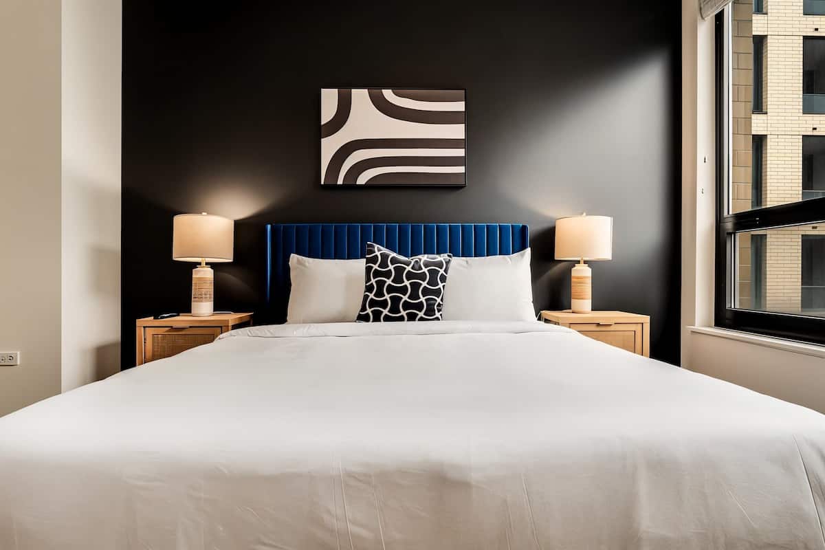 A modern hotel room with a large bed, two bedside tables with lamps, a dark wall with a geometric artwork, and a window with a view of the building.