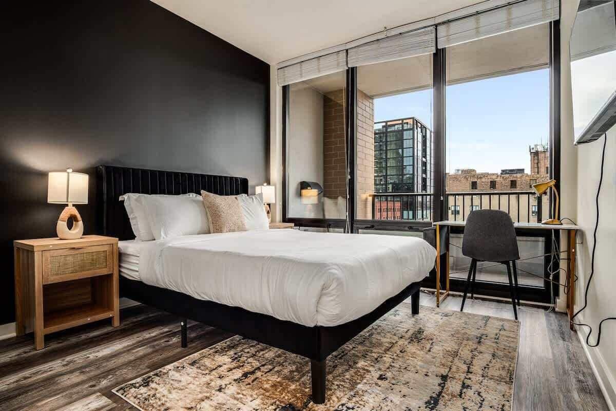 A modern hotel room with a large bed, a desk, and a view of the city through large windows.