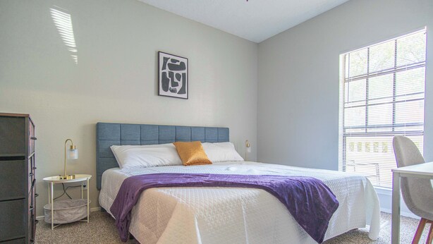 A bedroom with a bed, a nightstand, a chair, a desk, and a window. - Irving Apartments (Irving)