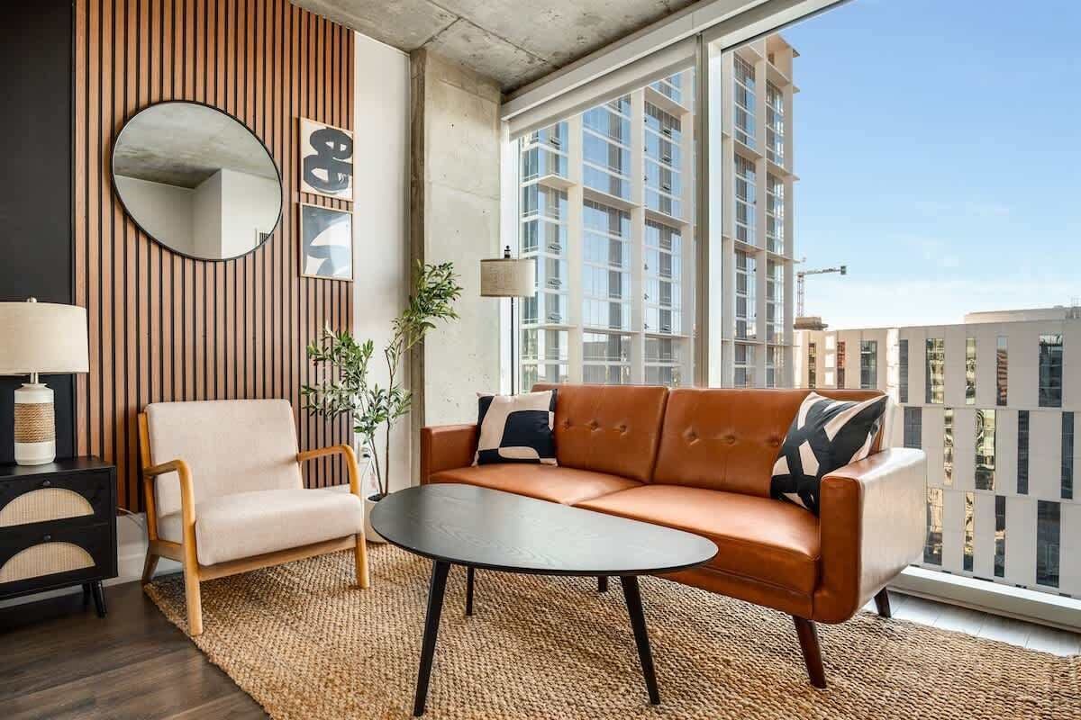 Una sala de estar moderna con sofá, sillón y mesa de centro, ventanas amplias y vista a la ciudad.
