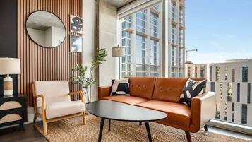 Una sala de estar moderna con sofá, sillón y mesa de centro, ventanas amplias y vista a la ciudad.