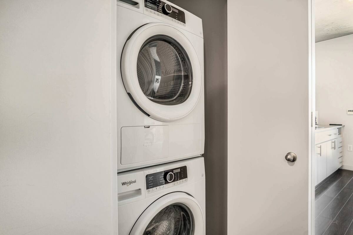 A stacked washer and dryer unit in a room with a door leading to a bathroom with a white cabinet and dark tiled floor.