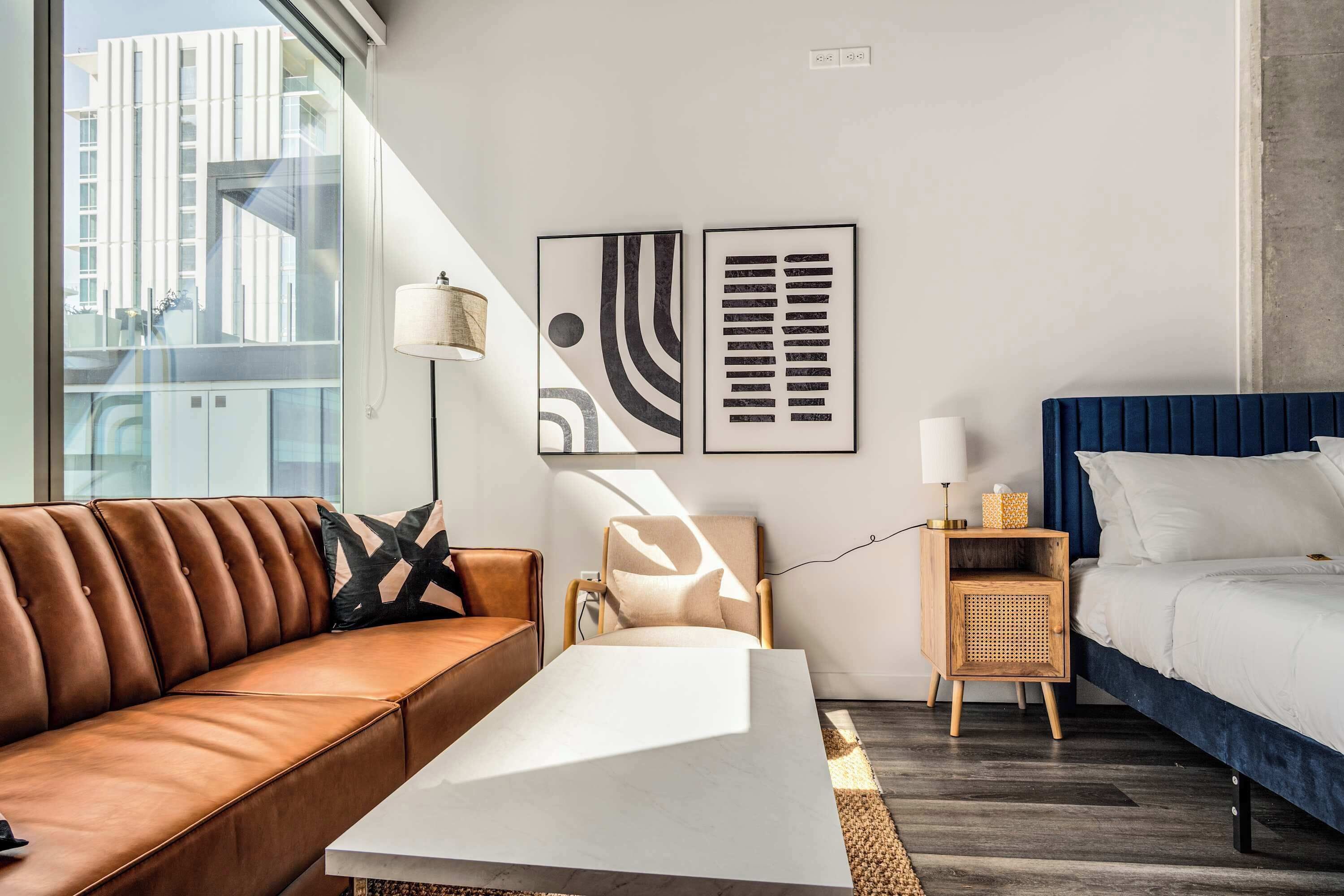 A modern hotel room with a leather sofa, a white coffee table, a blue bed, and two abstract wall art pieces.