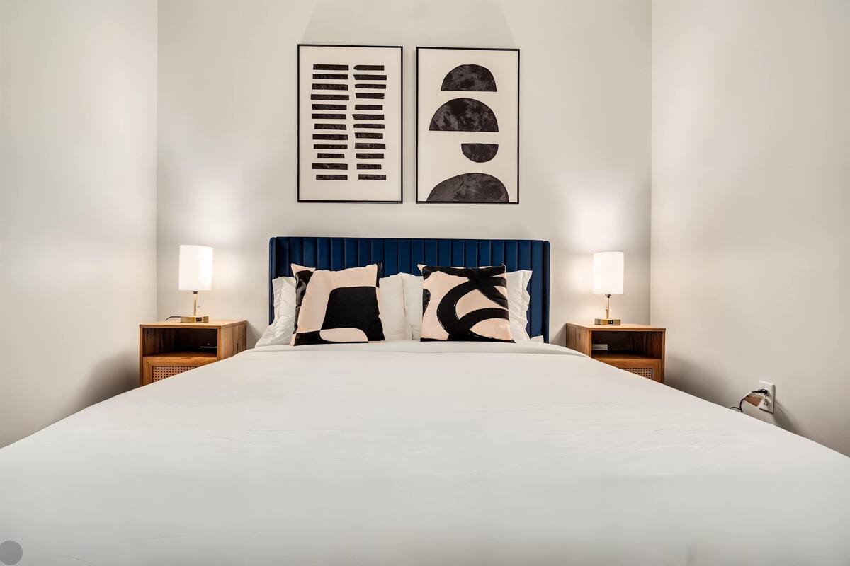 A neatly made bed with a blue headboard, two bedside tables with lamps, and two framed artworks on the wall.