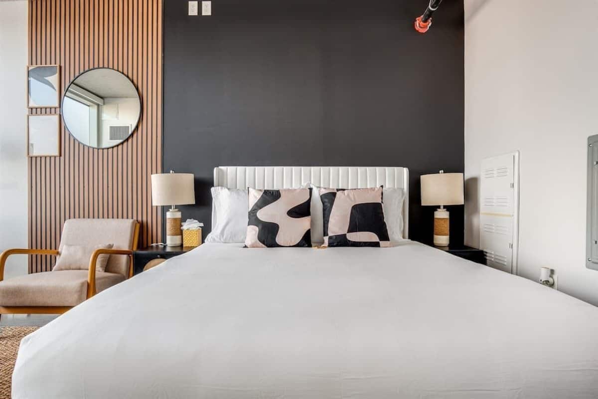 A hotel room with a large bed, two bedside lamps, a round mirror, a wooden panel wall, and a chair.