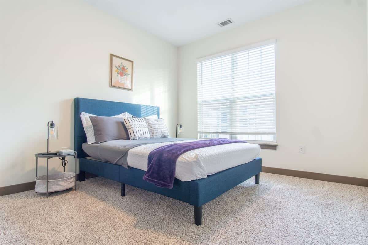 Une chambre à coucher comprenant un lit, une table de chevet, une lampe, une fenêtre avec des stores et un tableau accroché au mur.