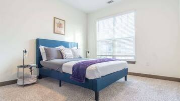 Une chambre à coucher comprenant un lit, une table de chevet, une lampe, une fenêtre avec des stores et un tableau accroché au mur.