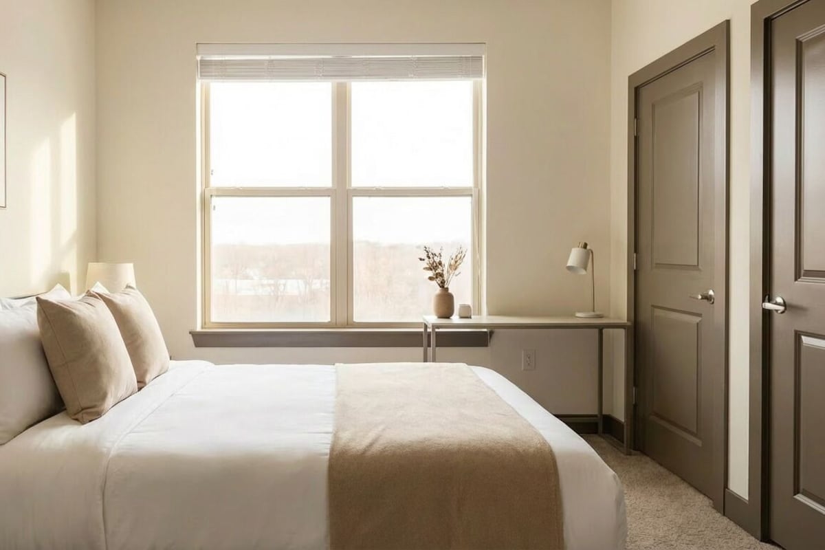 Un dormitorio con cama, mesita de noche, lámpara, ventana y puerta.