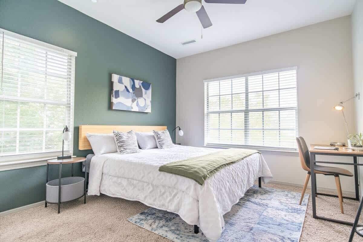 A bedroom with a bed, a desk, and a chair. There are two windows and a ceiling fan.