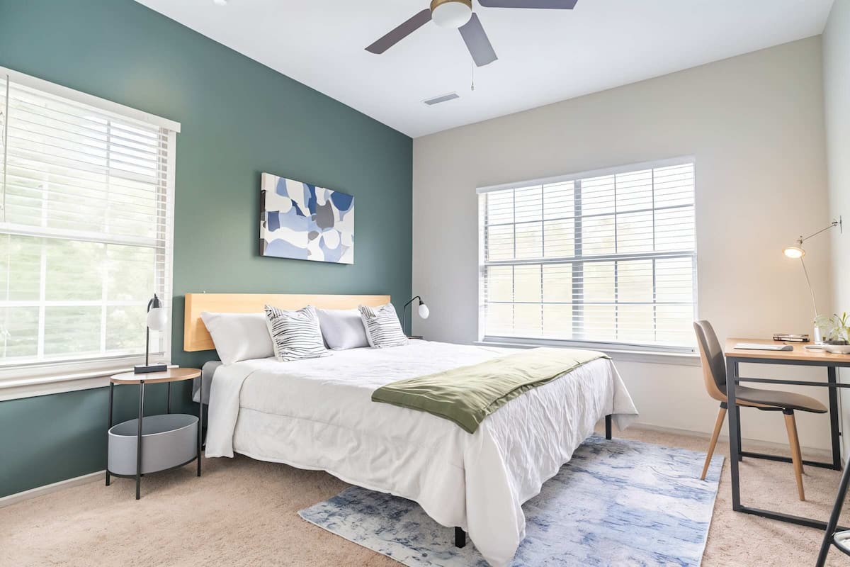 A bedroom with a bed, bedside table, lamp, and a desk with a chair.