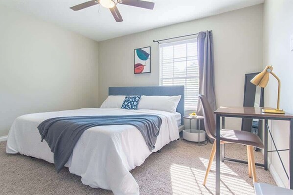 A bedroom with a bed, a desk, a chair, a ceiling fan, a window with curtains, and a wall-mounted artwork. - Boulevard Stays - North Dallas (Dallas)
