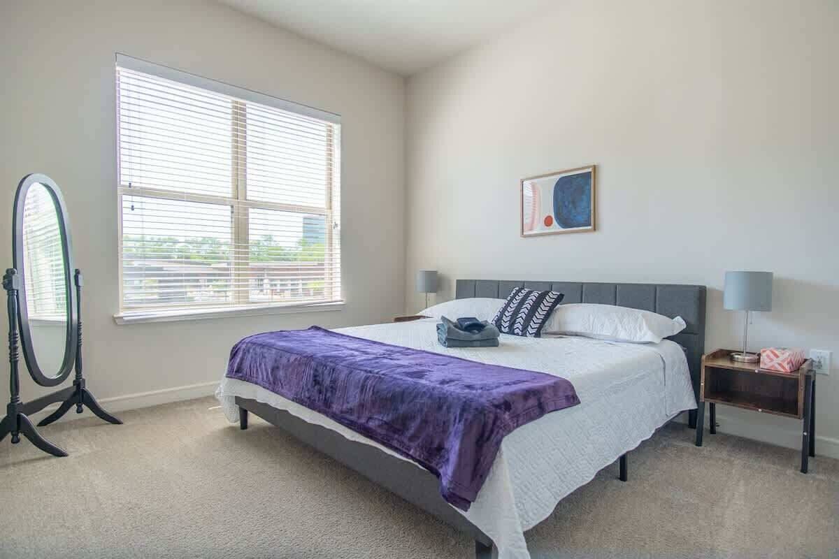 Un dormitorio con cama, una ventana con persianas, una mesita de noche con lámpara y una obra de arte colgada en la pared.
