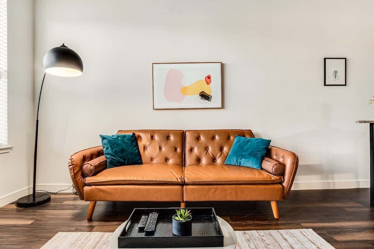 A brown leather sofa with teal cushions, a black floor lamp, and a framed abstract painting on the wall.