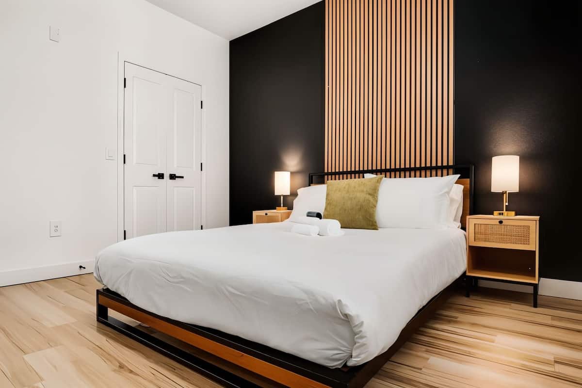 Une chambre d’hôtel moderne avec un grand lit, un plancher en bois, des murs noirs et blancs, et deux lampes de chevet.