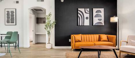 Una sala de estar moderna con un sofá, una mesa de centro y sillas. Hay una planta, una obra de arte en la pared y una lámpara.