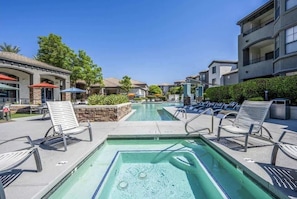 A pool area with lounge chairs, a clear pool, and buildings in the background.