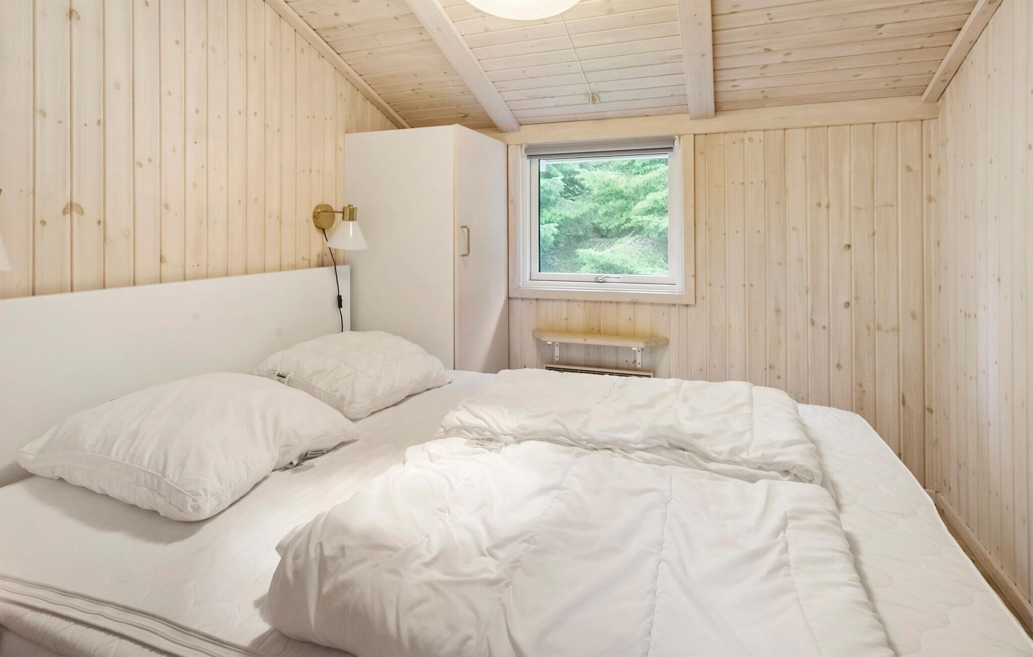A bedroom with a bed, white bedding, a nightstand, a lamp, a window with a view of greenery, and wooden walls.