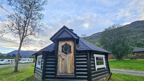 A log cabin with a hexagonal window and a door, surrounded by a grassy area and trees, with mountains in the background. - 2 bedroom lovely home in Røldal (Røldal)