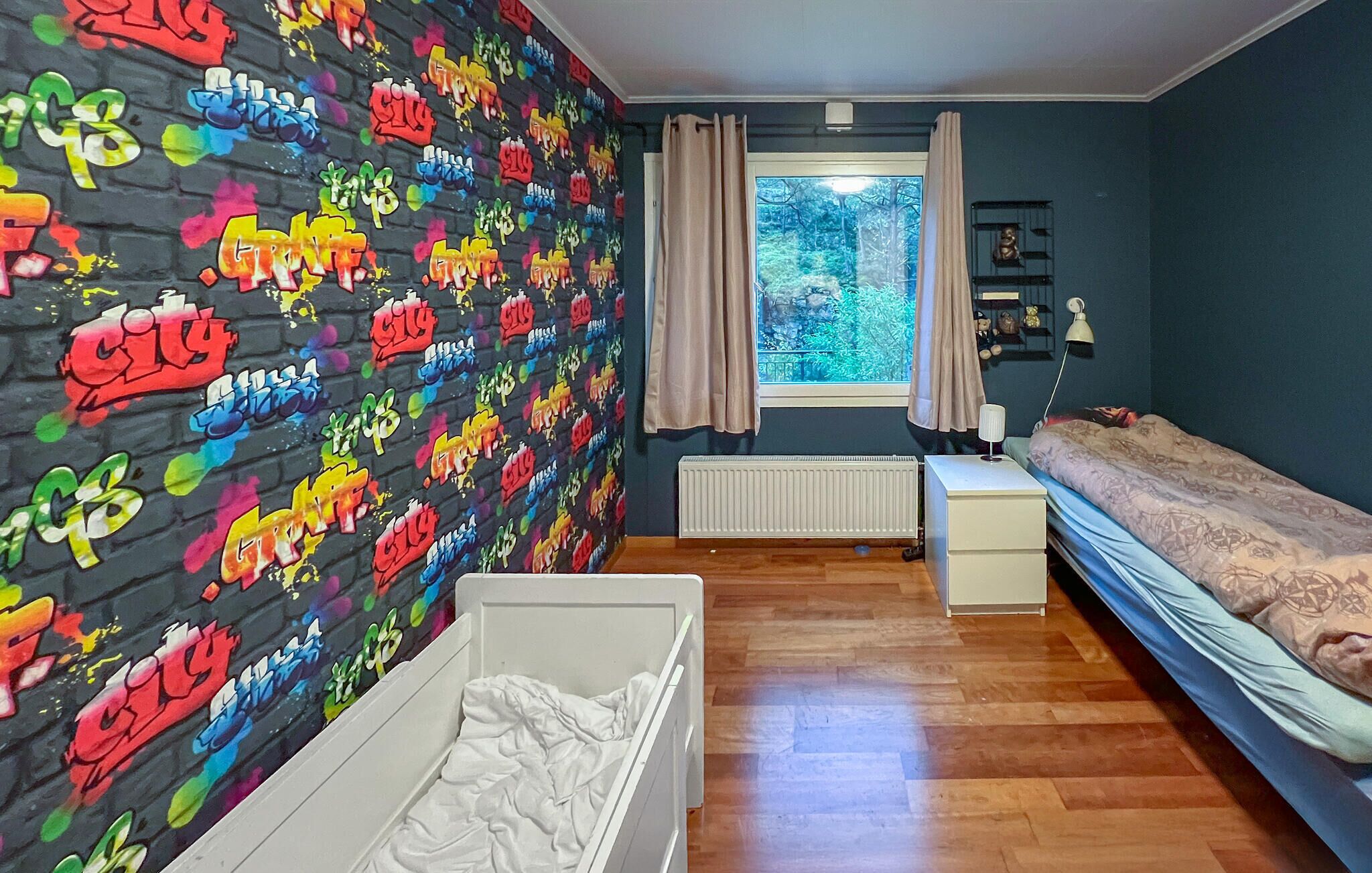Une chambre avec un mur décoré d’un motif en briques, une fenêtre avec des rideaux, un radiateur, une table de chevet blanche, un lit avec une couette à motifs et un meuble de rangement blanc.