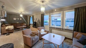 A cozy living room with a sofa, armchairs, a coffee table, and a TV. There are two windows with curtains, a chandelier, and a radiator. - Cozy home in Flekkefjord with house sea view (Flekkefjord)
