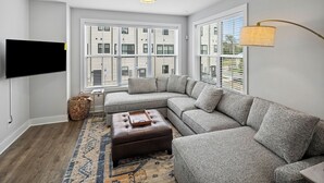 A modern living room with a sectional sofa, a flat-screen TV mounted on the wall, and a large window with blinds.