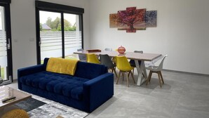 A modern living room with a blue sofa, a dining table with chairs, and a large window with blinds. - GITE DE L'ETOILE - ROCROI (ROCROI)