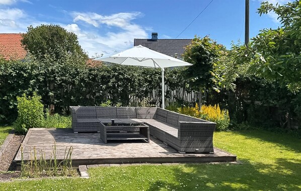 A patio with a sectional sofa and a table, surrounded by a garden and a white umbrella.