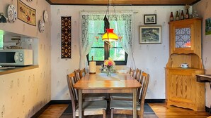 A dining area with a wooden table and chairs, a hanging light fixture, and a window with lace curtains.