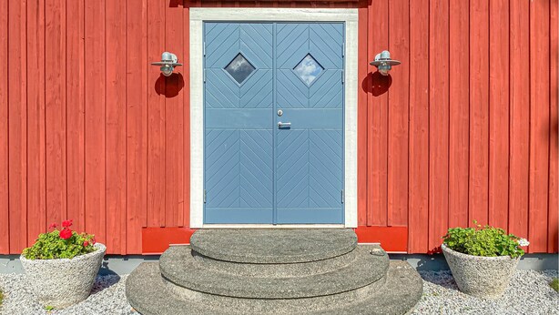 Un bâtiment rouge avec une porte bleue, deux lampes fixées au mur et deux plantes en pot.