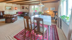 Un vaste salon comprenant un canapé en cuir, une table à manger avec des chaises et un tapis rouge à motifs. Les murs sont ornés de tableaux encadrés et une grande fenêtre est agrémentée de rideaux.