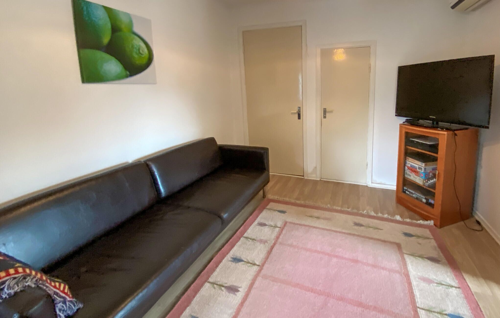 A living room with a black leather sofa, a pink rug, a television on a wooden stand, and a door in the background.