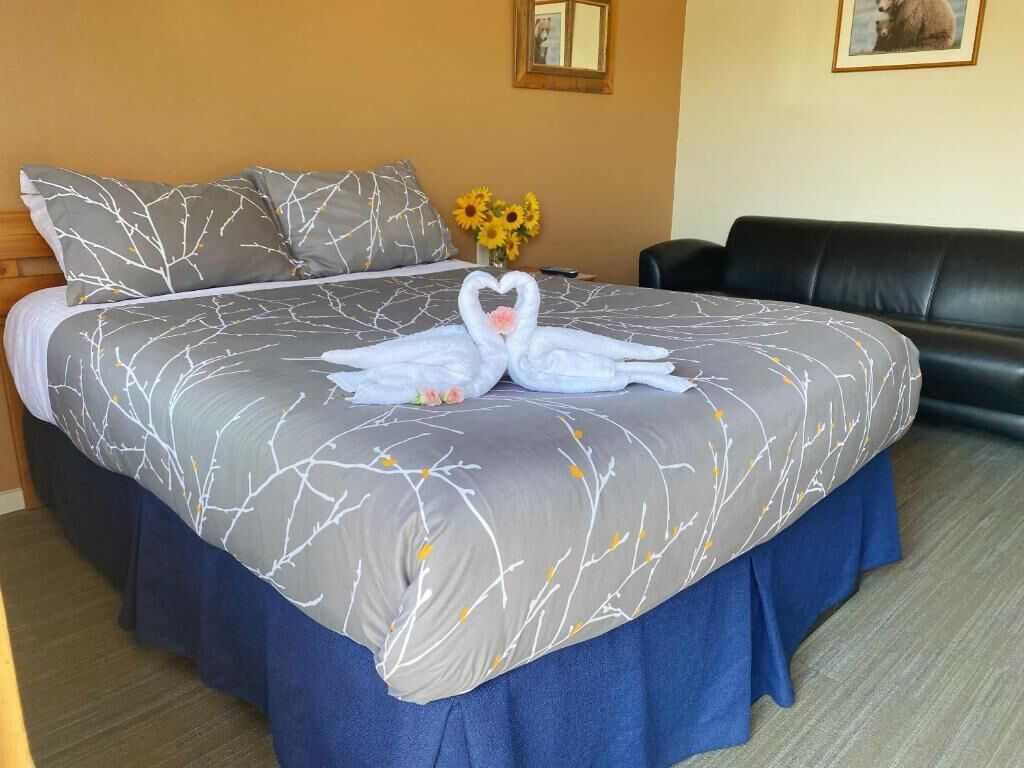 A neatly made bed with a grey and white tree branch pattern bedspread, a blue skirted bed base, and a heart-shaped towel arrangement on the bed.