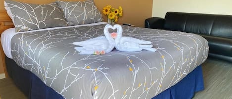 A neatly made bed with a grey and white tree branch pattern bedspread, a blue skirted bed base, and a heart-shaped towel arrangement on the bed.