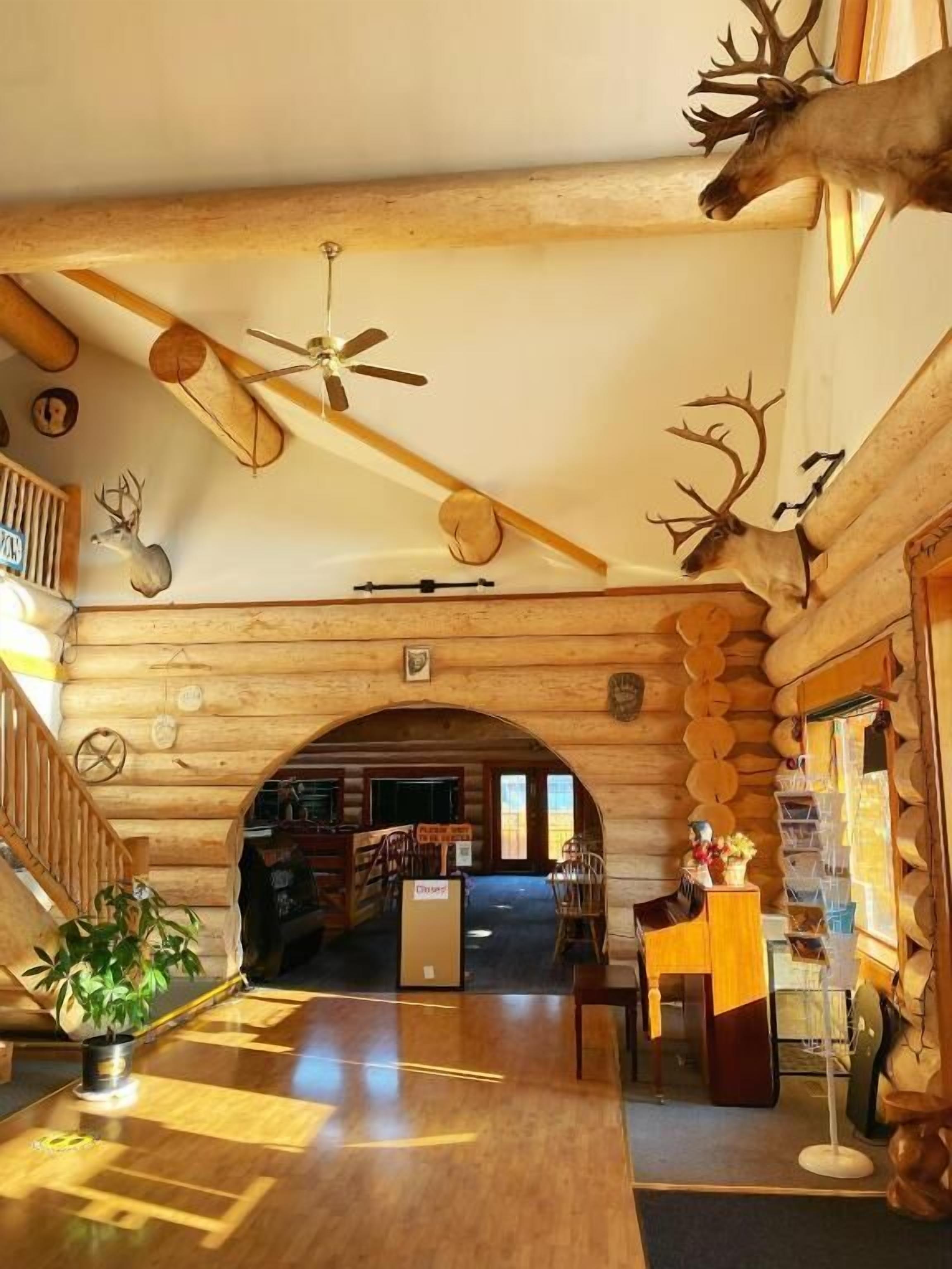 A log cabin interior with wooden walls, a wooden floor, a ceiling fan, and deer antlers mounted on the walls.