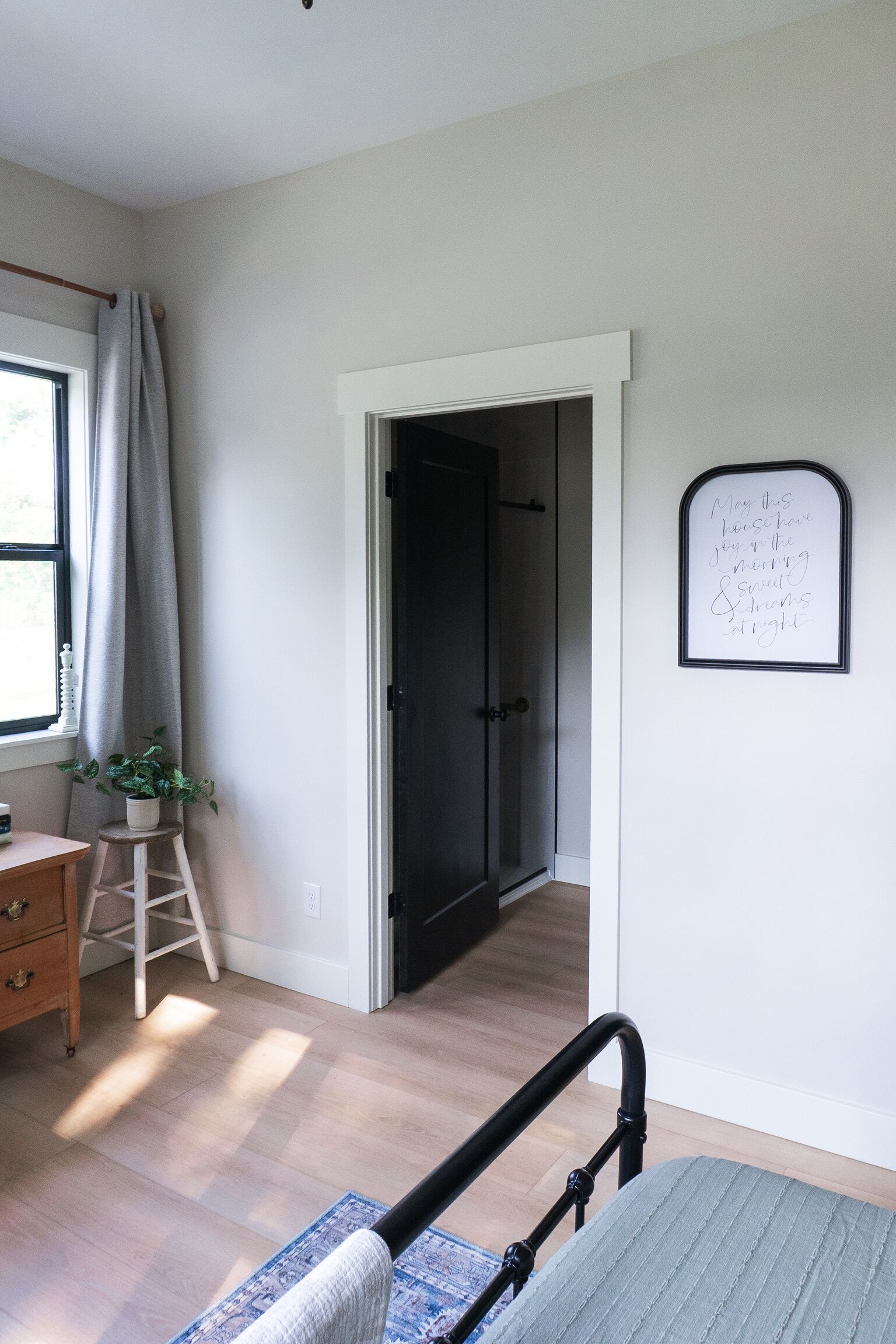 A room with a bed, a desk, a window with curtains, a door, and a wall with a framed quote.