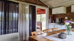 Een houten eettafel met stoelen, een houten vloer, een keuken met een fornuis en een spoelbak, en uitzicht op de omgeving door een raam met gordijnen.