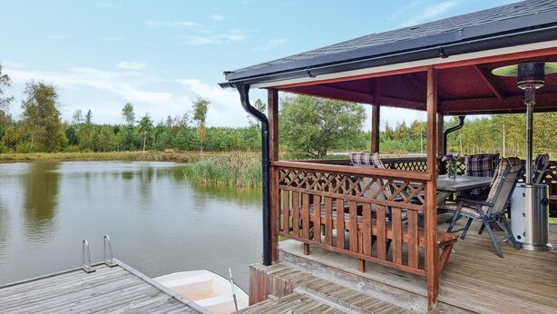 Een houten terras met een overdekte zithoek met uitzicht op een rustig meer, omringd door bomen.