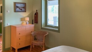 A bedroom with a wooden dresser, a wicker chair, a bed with a white duvet, a window with blinds, and a painting on the wall. - Pet friendly home in Vätö with sauna (Vätö)